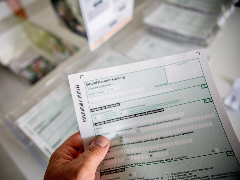 Nahaufnahme einer Person, die ein Formular in der Hand hält. Im Hintergrund unscharf ist ein Schreibtisch zu sehen, auf dem weitere Formulare liegen.