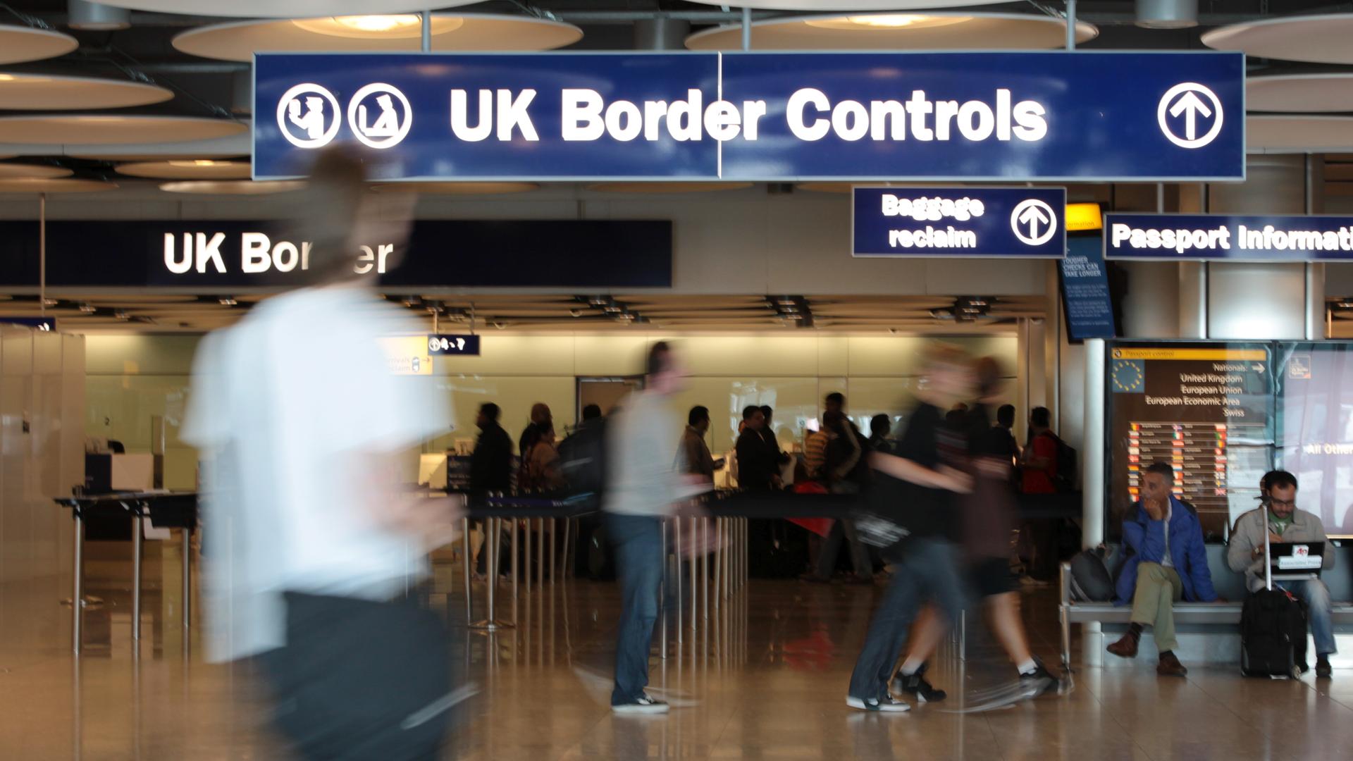 Blick auf den Gang mit dem Schild "UK Border Controls". Davor gehen Menschen, die man nur unscharf sieht. 