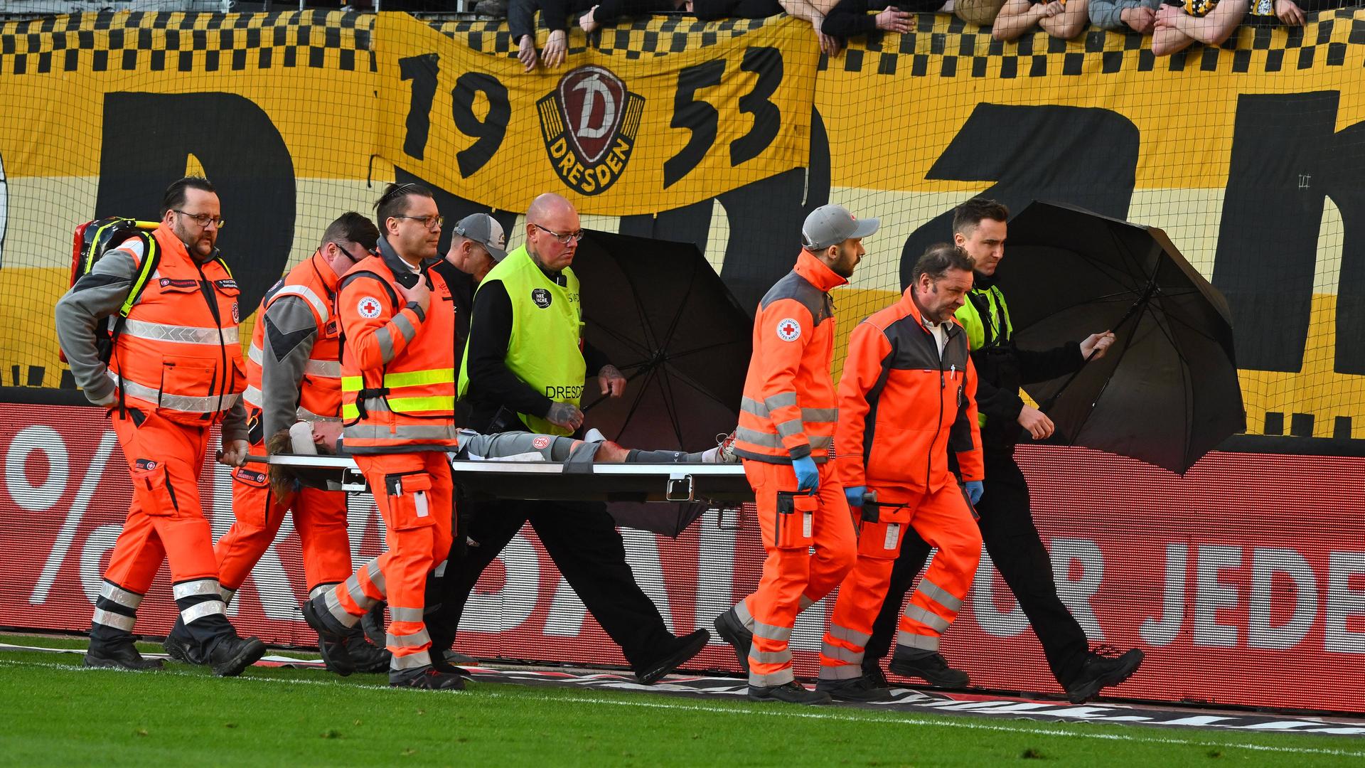 Sanitäter bringen einen verletzten Fußballer mit einer Trage vom Platz, geschützt gegen Wurfgeschosse von Ordnern mit Regenschirmen. Sanitäter bringen einen verletzten Fußballer mit einer Trage vom Platz, geschützt gegen Wurfgeschosse von Ordnern mit Regenschirmen.