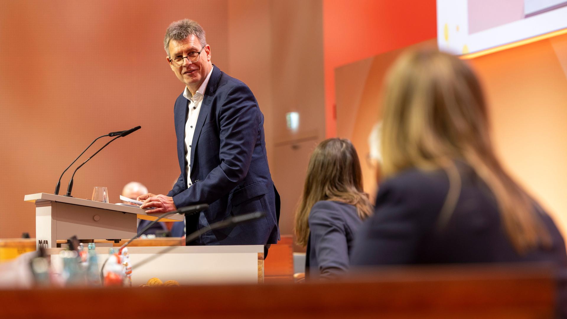 DOSB Präsident Thomas Weikert steht in einem blauen Anzug am Rednerpult. Er schaut zur Seite und lächelt.