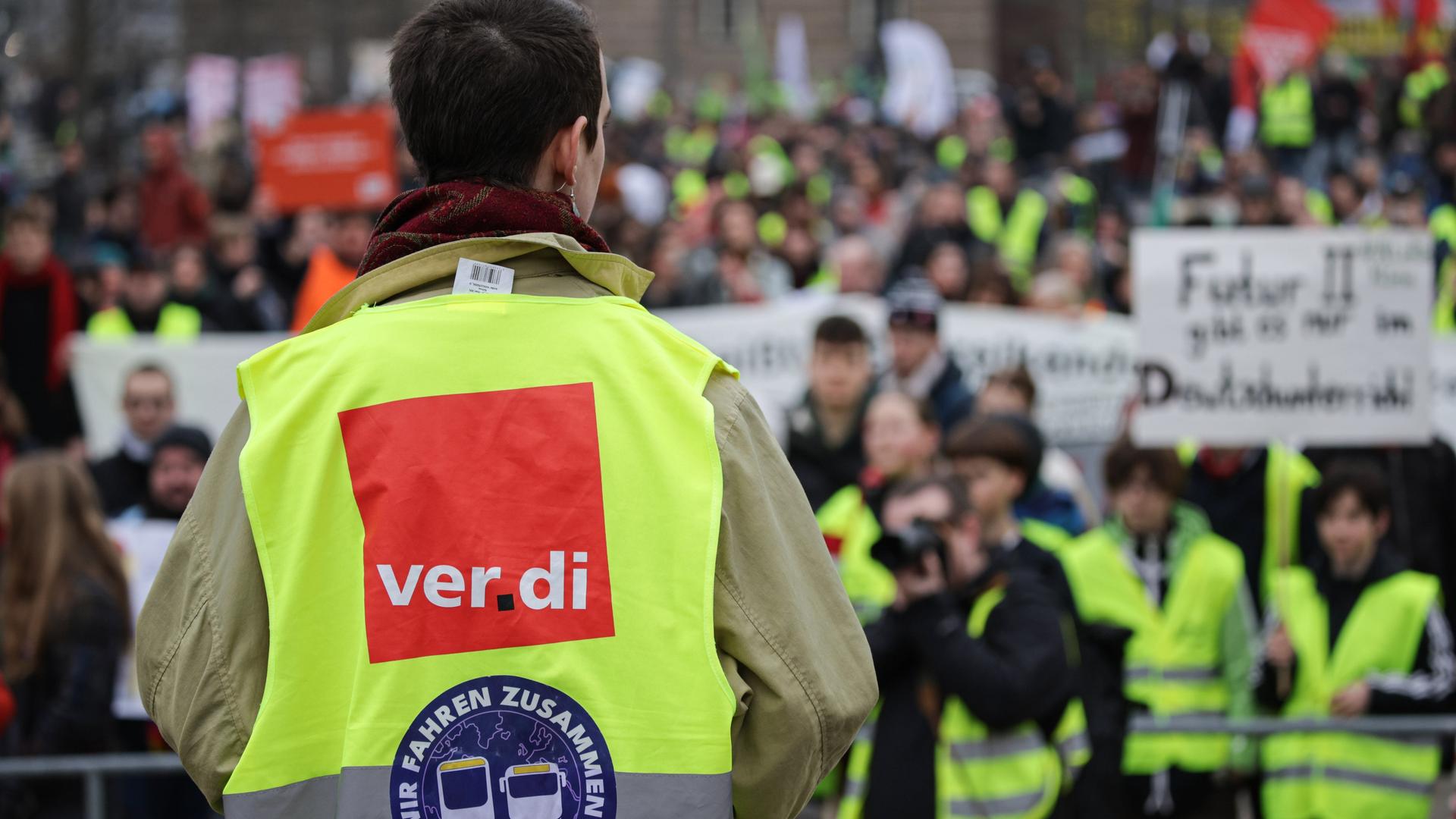 Ein Mann mit einer Verdi-Weste steht mit dem Rücken zu einer größeren Menschenmenge aus Streikenden.