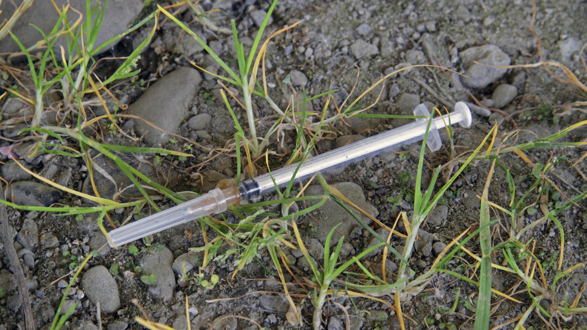 Eine Heroinspritze liegt auf sandigem Untergrund, umgeben von Grasbüscheln.