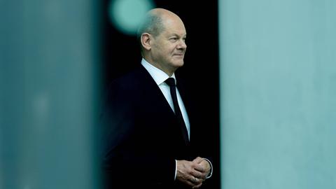 Olaf Scholz trägt einen schwarzen Anzug und steht zwischen zwei grauen Säulen, die in der Unschärfe zu erkennen sind