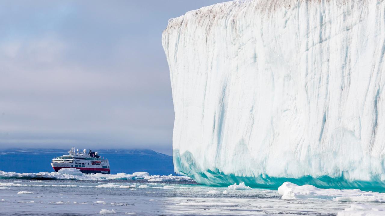 Ein Kreuzfahrtschiff fährt in der Arktis an einem gewaltigen weißen Eisberg vorbei. Im Hintergrund ragen schroffe, schneebedeckte Berge auf.