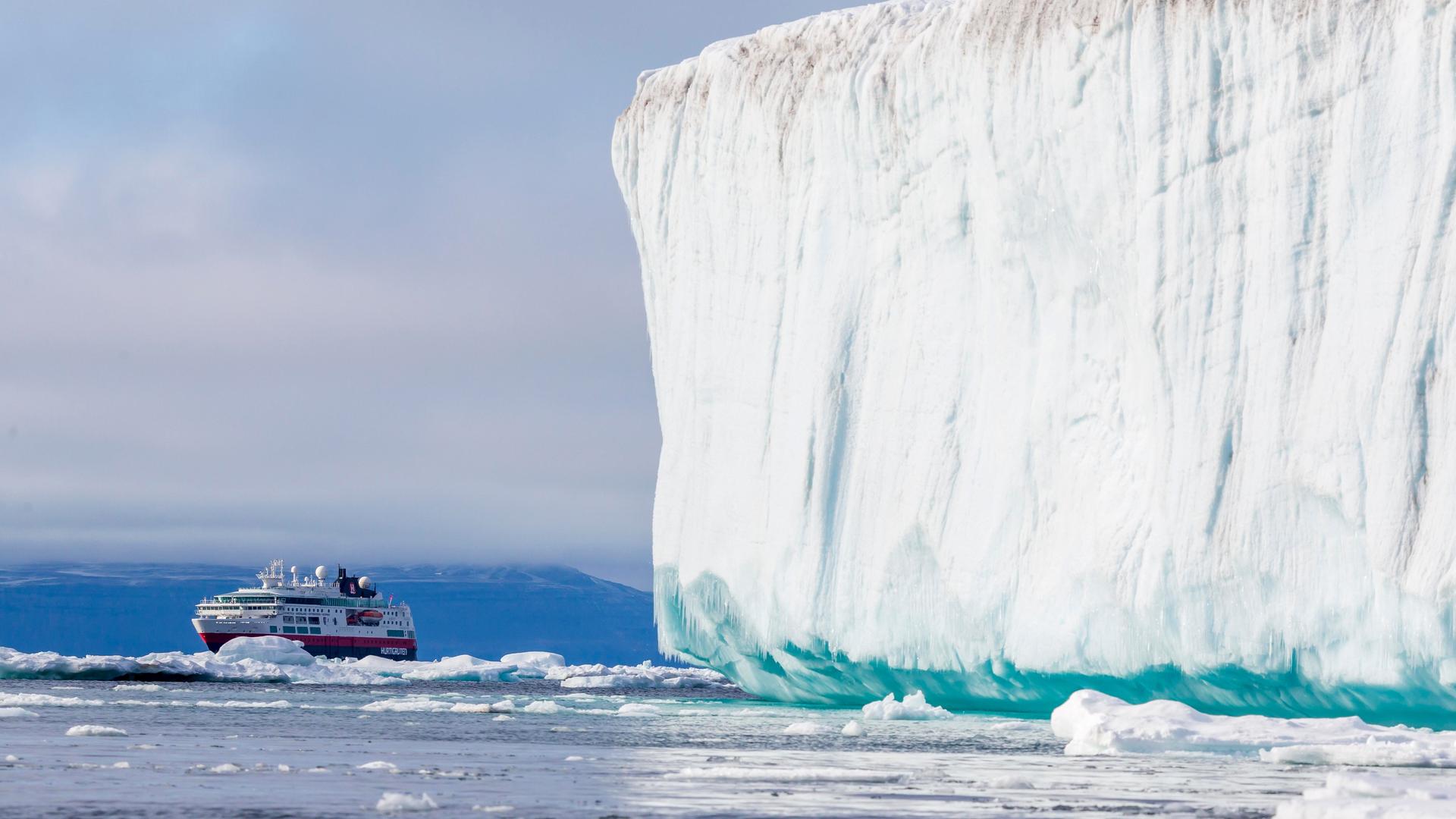 Ein Kreuzfahrtschiff fährt in der Arktis an einem gewaltigen weißen Eisberg vorbei. Im Hintergrund ragen schroffe, schneebedeckte Berge auf.