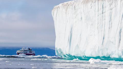 Ein Kreuzfahrtschiff fährt in der Arktis an einem gewaltigen weißen Eisberg vorbei. Im Hintergrund ragen schroffe, schneebedeckte Berge auf.