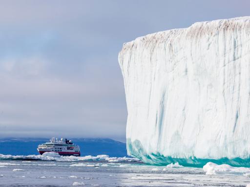 Ein Kreuzfahrtschiff fährt in der Arktis an einem gewaltigen weißen Eisberg vorbei. Im Hintergrund ragen schroffe, schneebedeckte Berge auf.