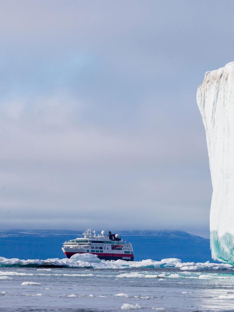 Ein Kreuzfahrtschiff fährt in der Arktis an einem gewaltigen weißen Eisberg vorbei. Im Hintergrund ragen schroffe, schneebedeckte Berge auf. Ein Kreuzfahrtschiff fährt in der Arktis an einem gewaltigen weißen Eisberg vorbei. Im Hintergrund ragen schroffe, schneebedeckte Berge auf.