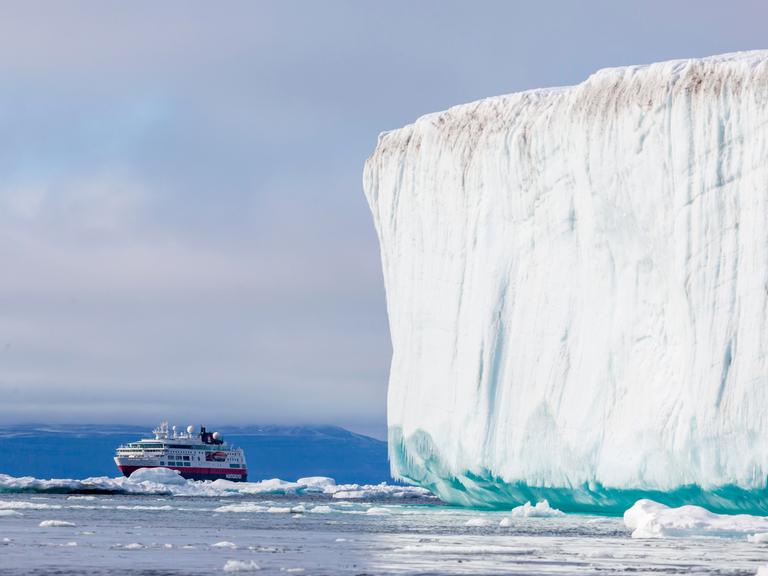 Ein Kreuzfahrtschiff fährt in der Arktis an einem gewaltigen weißen Eisberg vorbei. Im Hintergrund ragen schroffe, schneebedeckte Berge auf.
