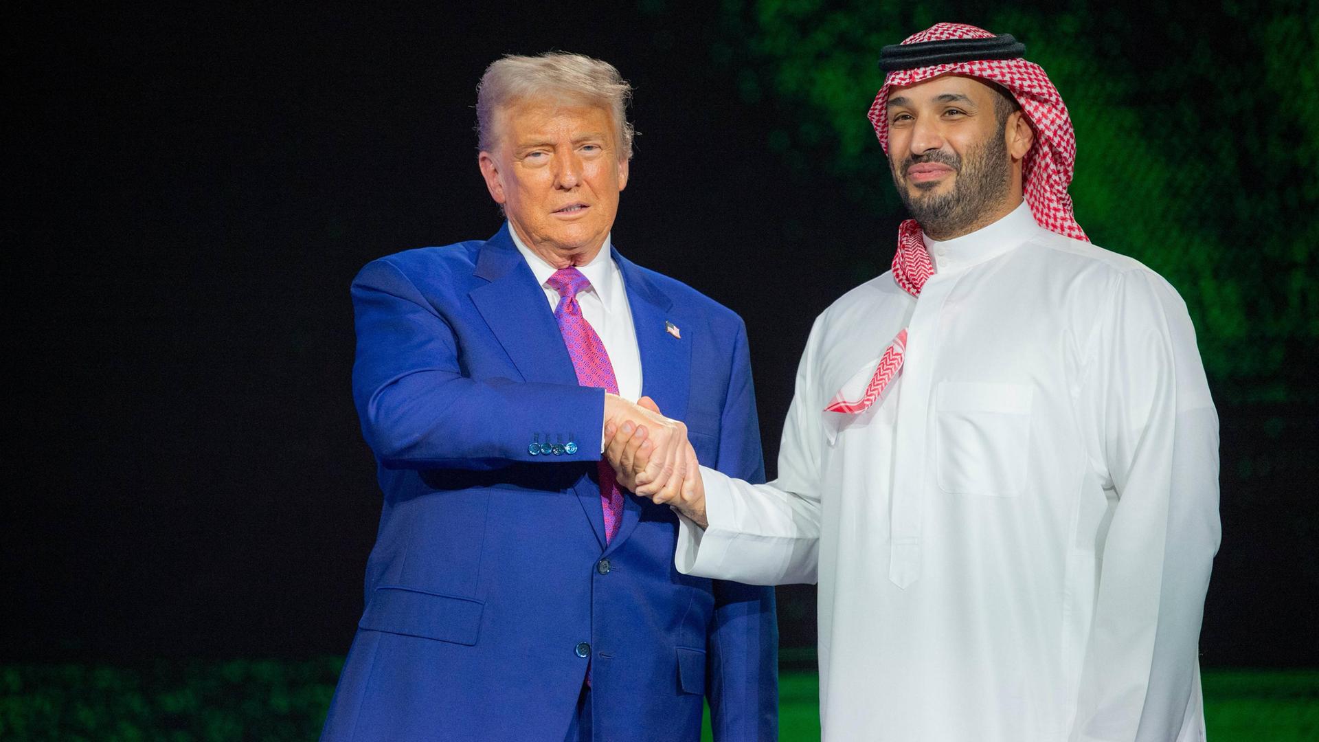 US-Präsident Trump in einem meerblauen Anzug schüttelt die Hand des saudischen Kronprinzen Mohammed bin Salman, der in traditionellem weissen Gewandt und einem Rot-weissen Ghutra auf dem Kopf auf der Bühne steht. US-Präsident Trump in einem meerblauen Anzug schüttelt die Hand des saudischen Kronprinzen Mohammed bin Salman, der in traditionellem weissen Gewandt und einem Rot-weissen Ghutra auf dem Kopf auf der Bühne steht.