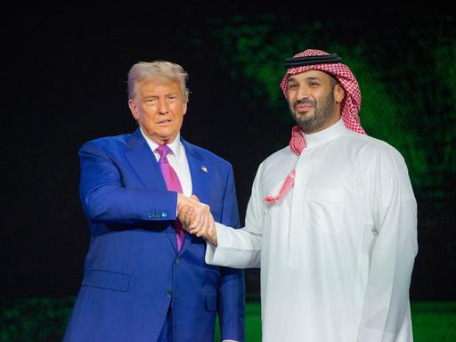 US-Präsident Trump in einem meerblauen Anzug schüttelt die Hand des saudischen Kronprinzen Mohammed bin Salman, der in traditionellem weissen Gewandt und einem Rot-weissen Ghutra auf dem Kopf auf der Bühne steht. 