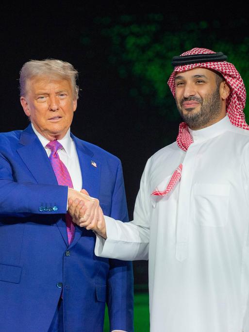 US-Präsident Trump in einem meerblauen Anzug schüttelt die Hand des saudischen Kronprinzen Mohammed bin Salman, der in traditionellem weissen Gewandt und einem Rot-weissen Ghutra auf dem Kopf auf der Bühne steht. 