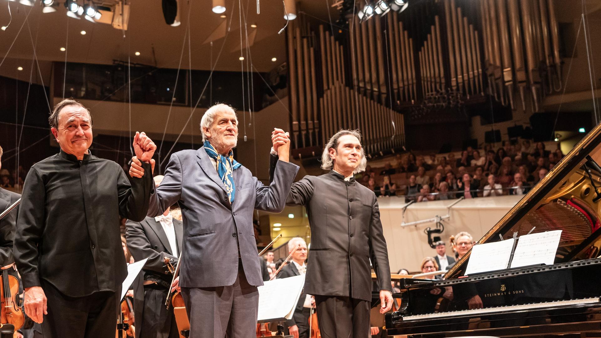 Drei Männer stehen auf der Bühne der Philharmonie und verbeugen sich. Drei Männer stehen auf der Bühne der Philharmonie und verbeugen sich.