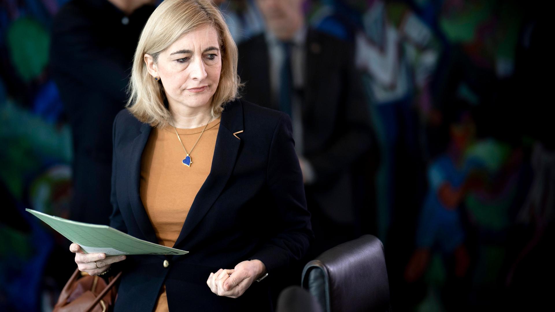 Eine Frau mit blonden Haaren und schwarzem Sakko hält eine Mappe in der Hand