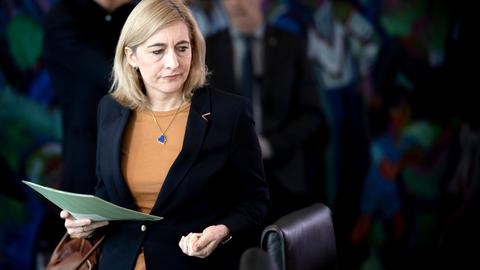 Eine Frau mit blonden Haaren und schwarzem Sakko hält eine Mappe in der Hand