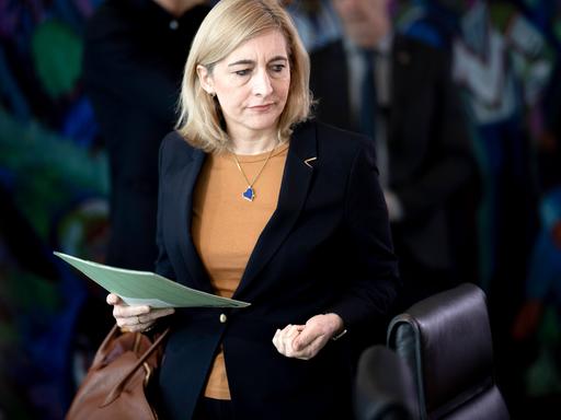 Eine Frau mit blonden Haaren und schwarzem Sakko hält eine Mappe in der Hand