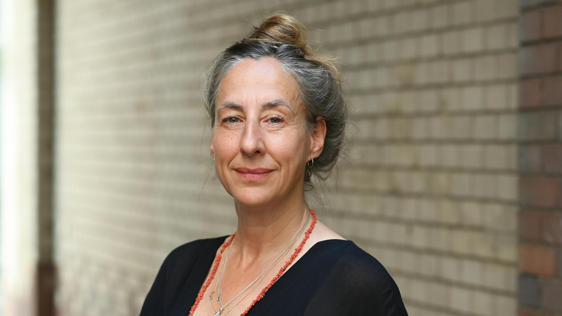 Die Schriftstellerin Judith Hermann steht vor einer Backsteinwand und blickt in die Kamera. Ihr angegrautes Haar hat sie hochgesteckt, sie trägt ein schwarzes Oberteil und zwei Ketten, eine davon rot.