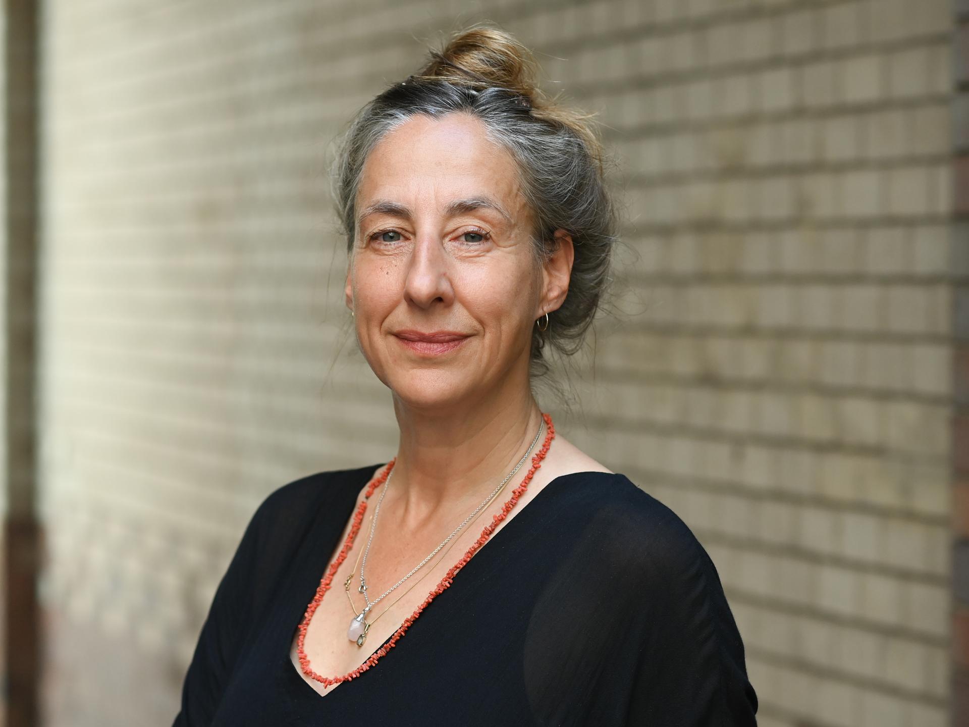 Die Schriftstellerin Judith Hermann steht vor einer Backsteinwand und blickt in die Kamera. Ihr angegrautes Haar hat sie hochgesteckt, sie trägt ein schwarzes Oberteil und zwei Ketten, eine davon rot.
