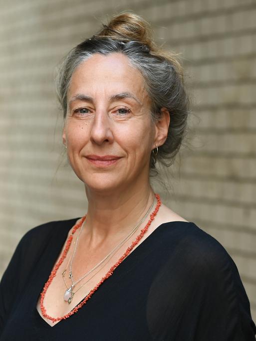 Die Schriftstellerin Judith Hermann steht vor einer Backsteinwand und blickt in die Kamera. Ihr angegrautes Haar hat sie hochgesteckt, sie trägt ein schwarzes Oberteil und zwei Ketten, eine davon rot.