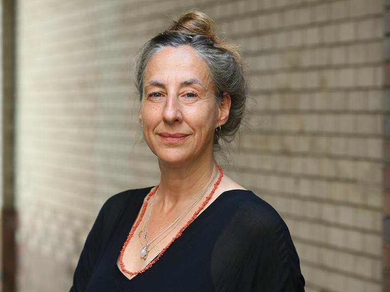Die Schriftstellerin Judith Hermann steht vor einer Backsteinwand und blickt in die Kamera. Ihr angegrautes Haar hat sie hochgesteckt, sie trägt ein schwarzes Oberteil und zwei Ketten, eine davon rot.