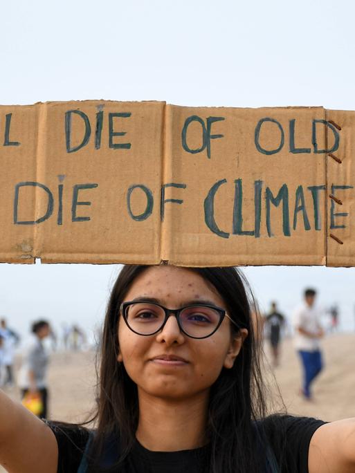 "Ihr sterbt an Altersschwäche, wir am Klimawandel": Eine junge Protestierende macht ihrem Ärger über zögerlich umgesetzte Klimaziele Luft, indem sie ein selbst beschriebenes Pappschild hochhält. Sie lächelt in die Kamera. 