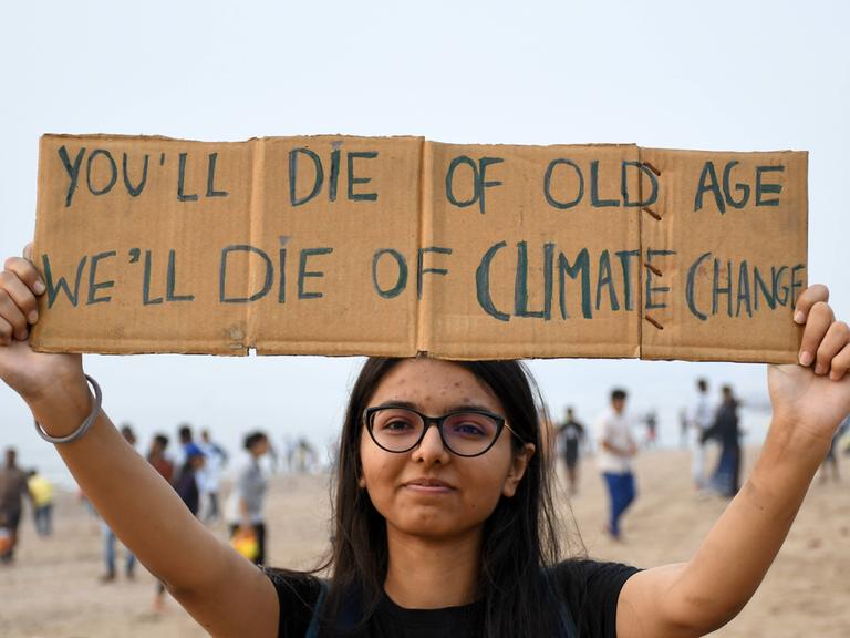 "Ihr sterbt an Altersschwäche, wir am Klimawandel": Eine junge Protestierende macht ihrem Ärger über zögerlich umgesetzte Klimaziele Luft, indem sie ein selbst beschriebenes Pappschild hochhält. Sie lächelt in die Kamera. 