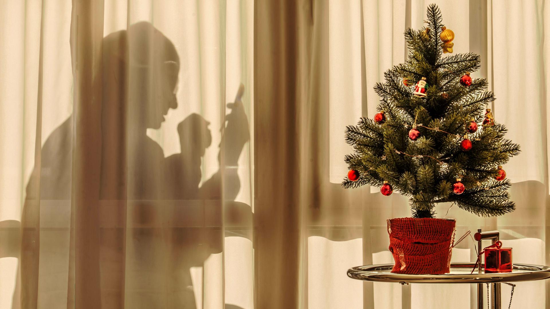 Ein älterer Mann sitzt an Weihnachten hinter einem transparenten Vorhang allein zuhause und schaut in sein Handy. Vor dem Vorhang steht ein kleiner geschmückter Weihnachtsbaum.