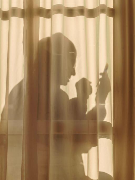 Ein älterer Mann sitzt an Weihnachten hinter einem transparenten Vorhang allein zuhause und schaut in sein Handy. Vor dem Vorhang steht ein kleiner geschmückter Weihnachtsbaum.