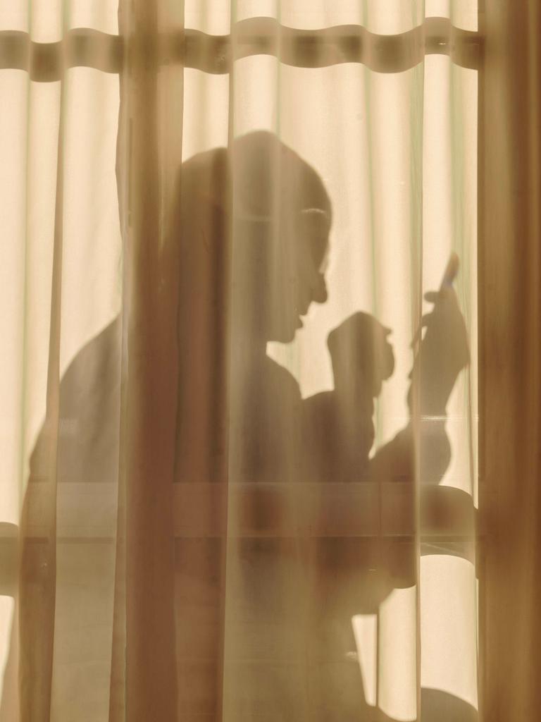 Ein älterer Mann sitzt an Weihnachten hinter einem transparenten Vorhang allein zuhause und schaut in sein Handy. Vor dem Vorhang steht ein kleiner geschmückter Weihnachtsbaum.