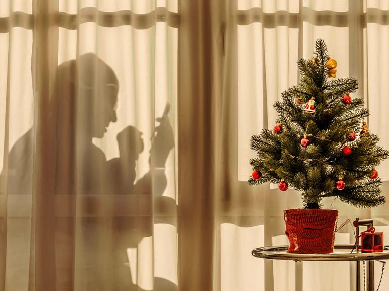 Ein älterer Mann sitzt an Weihnachten hinter einem transparenten Vorhang allein zuhause und schaut in sein Handy. Vor dem Vorhang steht ein kleiner geschmückter Weihnachtsbaum.