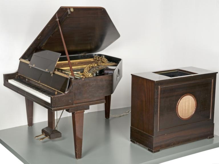 Blick auf einen holzfarbenen Flügel, der mit einer großen Lautsprecher-Box mit Drähten verbunden ist.