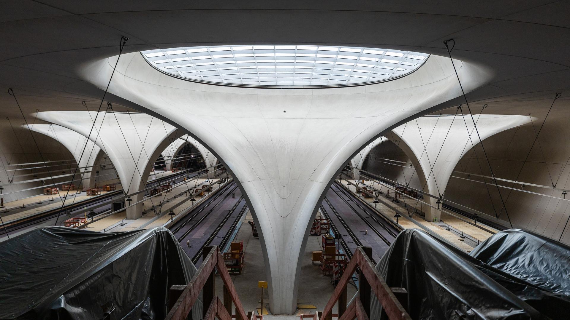 Blick in die Bahnhofshalle des zukünftigen Stuttgarter Hauptbahnhofs. Im Zentrum ein Betonelement, welches sich nach oben zu einem Glasfenster öffnet. Links und Rechts in Plane gehüllte Rolltreppen.