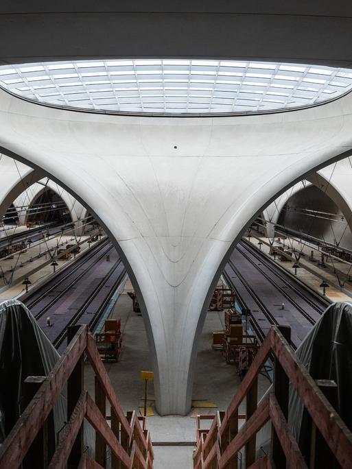 Blick in die Bahnhofshalle des zukünftigen Stuttgarter Hauptbahnhofs. Im Zentrum ein Betonelement, welches sich nach oben zu einem Glasfenster öffnet. Links und Rechts in Plane gehüllte Rolltreppen.