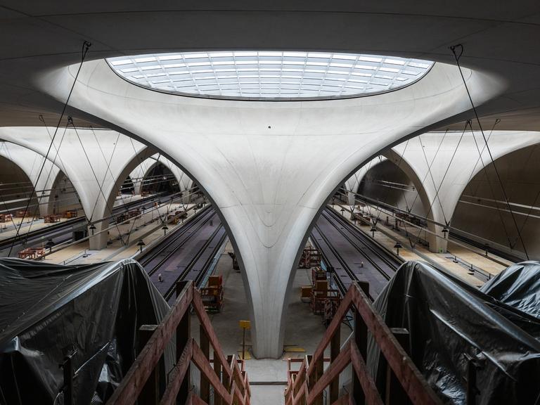 Blick in die Bahnhofshalle des zukünftigen Stuttgarter Hauptbahnhofs. Im Zentrum ein Betonelement, welches sich nach oben zu einem Glasfenster öffnet. Links und Rechts in Plane gehüllte Rolltreppen.
