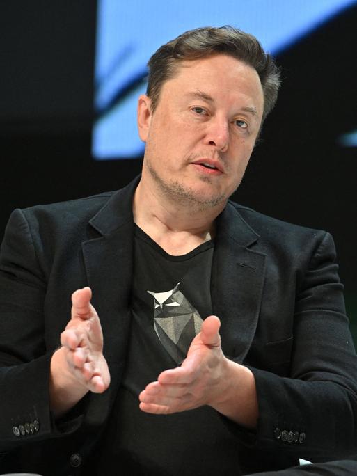 Elon Musk sitzt auf einem Stuhl und hält seine Hände mit offenen Handflächen vor sich