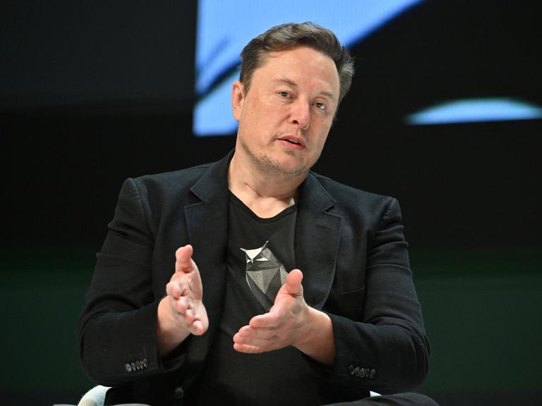 Elon Musk sitzt auf einem Stuhl und hält seine Hände mit offenen Handflächen vor sich