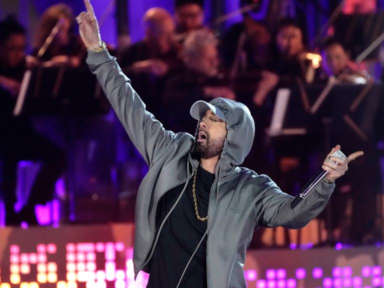 Ein Mann steht auf einer Bühne. Er hebt seine rechten Arm nach oben. Er trägt einen grauen Kapuzenpullover. der Sänger ist der US-Hiphoper Eminem.