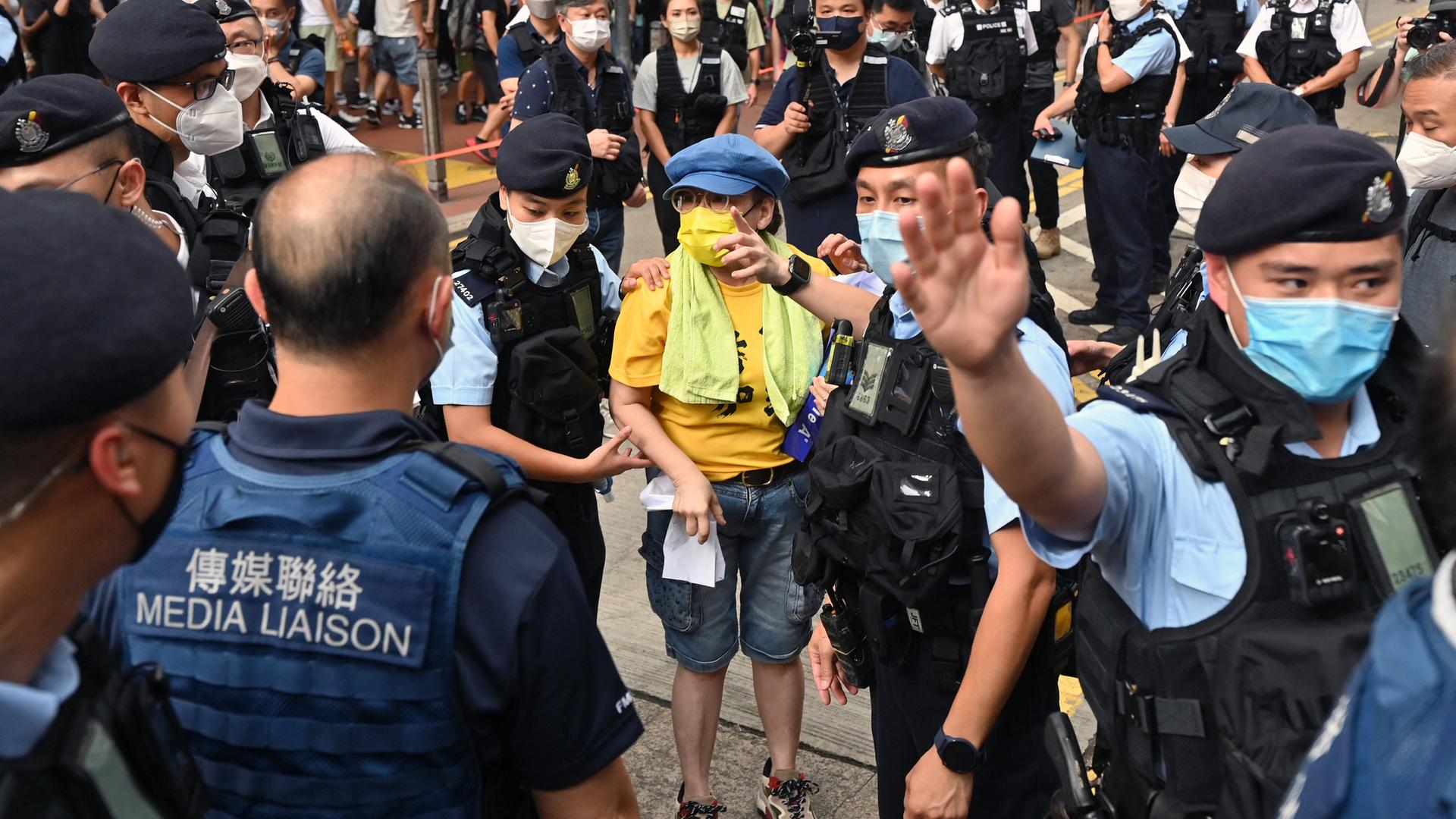 Zahlreiche Polizistinnen und Polizisten stehen um die Frau im gelben T-Shirt herum. Ein Polizist weist Passanten mit erhobenem Arm zurück. Zahlreiche Polizistinnen und Polizisten stehen um die Frau im gelben T-Shirt herum. Ein Polizist weist Passanten mit erhobenem Arm zurück.