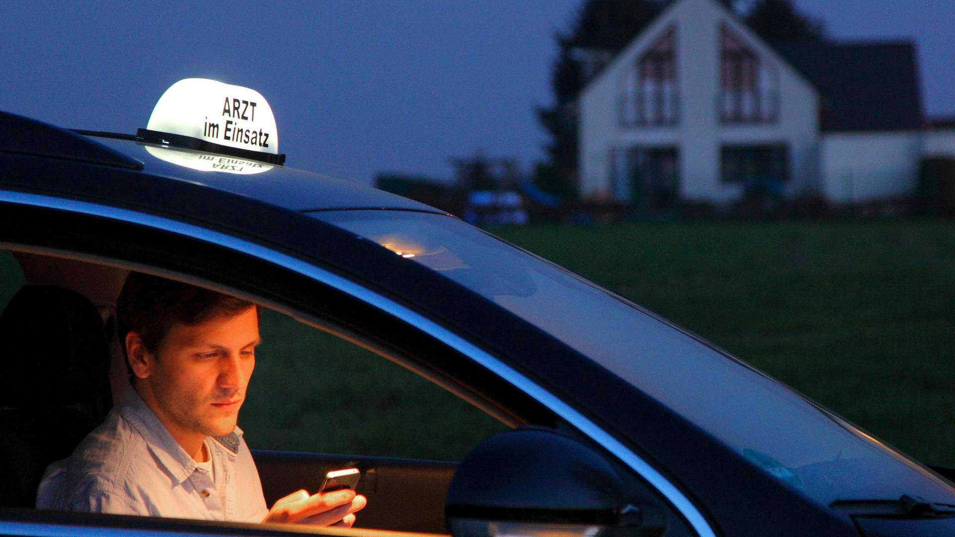 Ein junger Landarzt telefoniert nach einem abendlichen Hausbesuch in seinem Fahrzeug. Auf dem Dach seines Autos steht ein Schild mit der Aufschrift "Arzt im Einsatz"