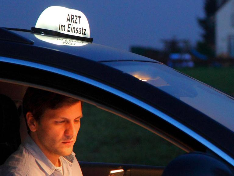 Ein junger Landarzt telefoniert nach einem abendlichen Hausbesuch in seinem Fahrzeug. Auf dem Dach seines Autos steht ein Schild mit der Aufschrift "Arzt im Einsatz"