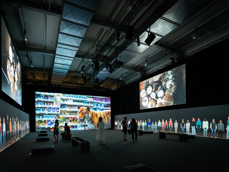 In einer Halle werden Bilder auf fünf Bildschirme projiziert. Das Zentralste zeigt einen Tiger vor einem Supermarktregal. Auf den seitlichen Monitoren sind ein Schlagzeuger und nebeneinander stehende Menschen in Lebensgröße zu sehen.