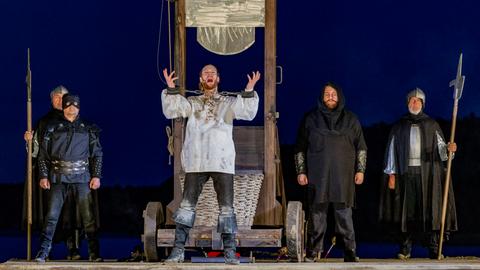 Premiere der Störtebeker-Festspiele 2024. Der Schauspieler Moritz Stephan (Mitte) als Klaus Störtebeker steht vor einer Guillotine und hebt die Hände. Er scheint laut zu sprechen.