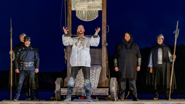 Premiere der Störtebeker-Festspiele 2024. Der Schauspieler Moritz Stephan (Mitte) als Klaus Störtebeker steht vor einer Guillotine und hebt die Hände. Er scheint laut zu sprechen.