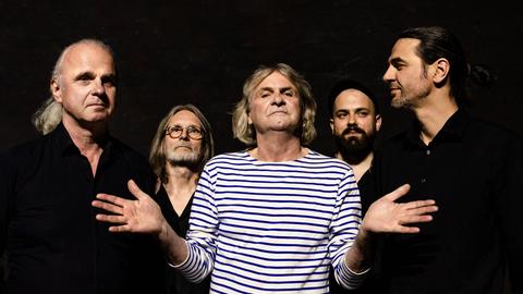 Ein Mann mit weiß-blaumen Ringelshirt steht mit ausgestreckten Händen vor der Kamera, umgeben von vier schwarz gekleideten Männern. Ein Mann mit weiß-blaumen Ringelshirt steht mit ausgestreckten Händen vor der Kamera, umgeben von vier schwarz gekleideten Männern.