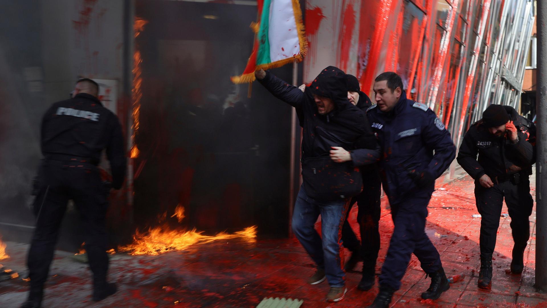 Polizisten verhaften einen Demonstranten. Im Hintergrund die EU-Vertretung in Sofia, vor der ein Feuer brennt.