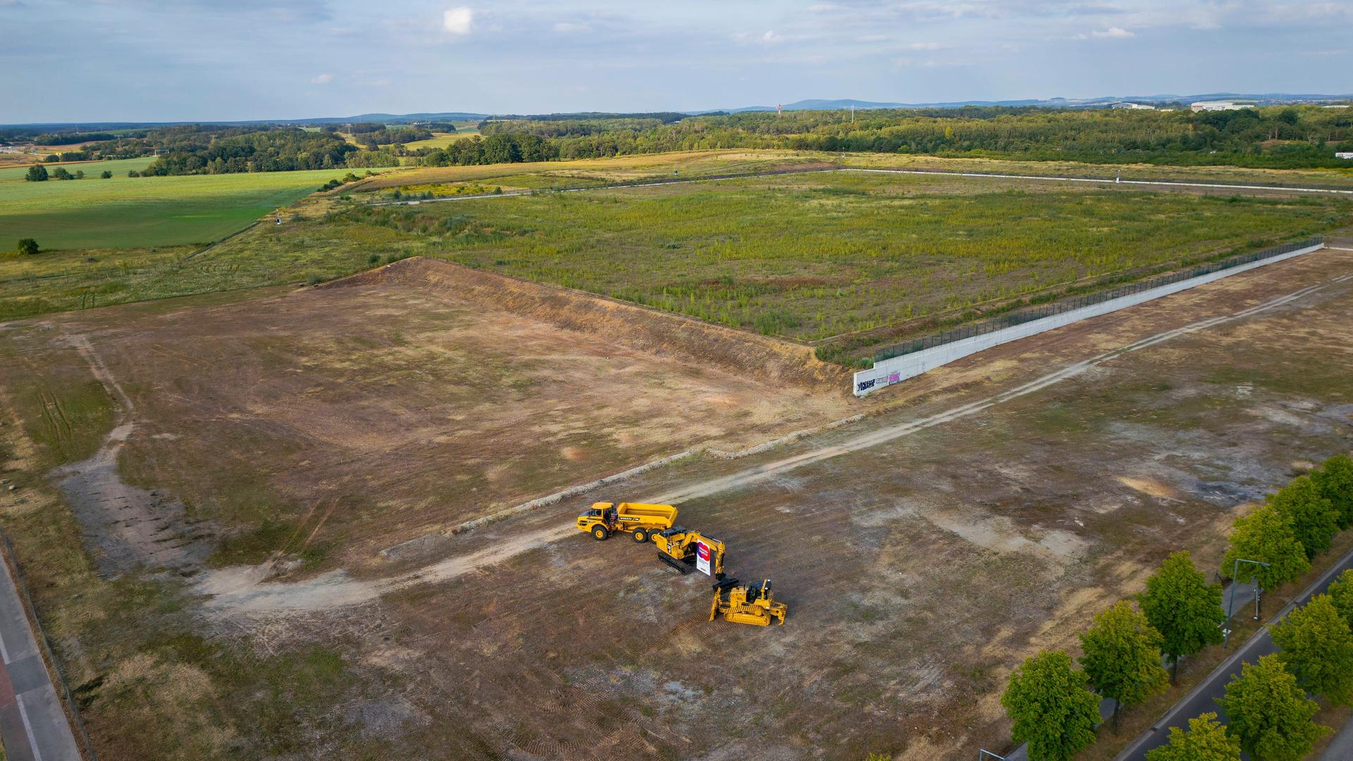 Hinter dem bewchten Bauzaun zeigen sich Vorzeichen des Baustartes. Baumaschienen stehen herum. Straßen, Wege und viel Wegetation kennzeichen die Ansicht.