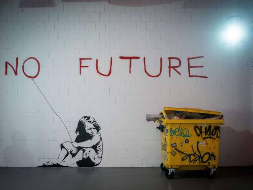 Ein großes Wandgraffiti zeigt den roten Schriftzug „no future“ und ein Mädchen, das daneben auf dem Boden kauert und das "o" wie einen Luftballon an einer Schnur hält. 