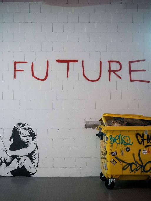 Ein großes Wandgraffiti zeigt den roten Schriftzug „no future“ und ein Mädchen, das daneben auf dem Boden kauert und das "o" wie einen Luftballon an einer Schnur hält. 