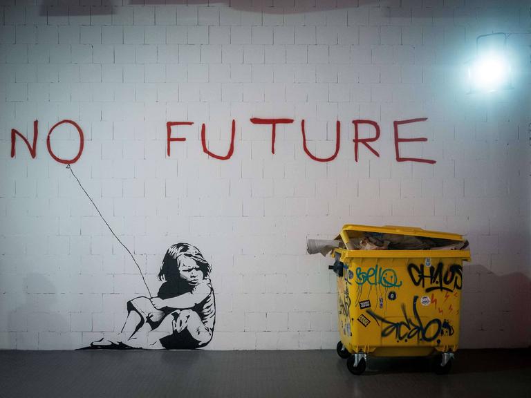 Ein großes Wandgraffiti zeigt den roten Schriftzug „no future“ und ein Mädchen, das daneben auf dem Boden kauert und das "o" wie einen Luftballon an einer Schnur hält. 