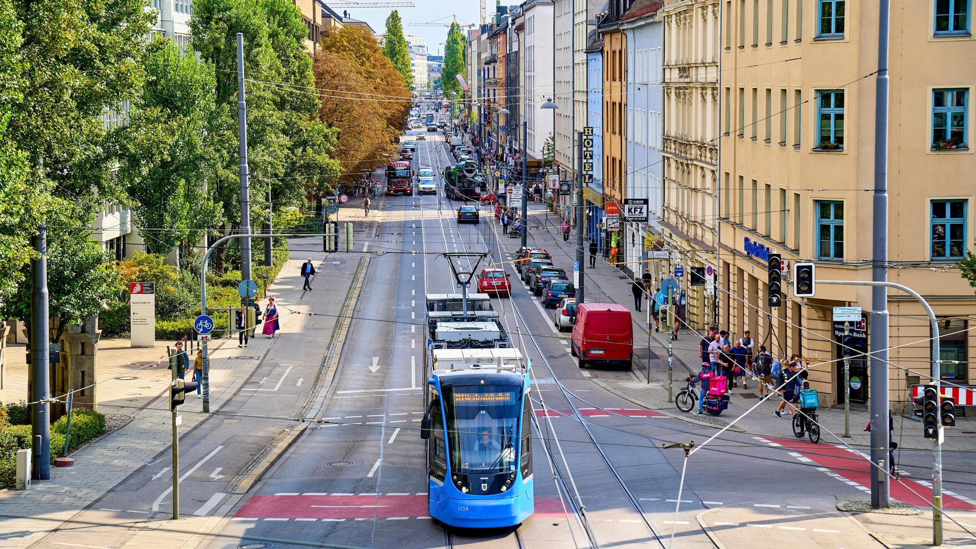 Eine blaue Straßenbahn fährt durch eine typische Straße in München, umgeben von geparkten Autos, Fußgängern, Radfahrern und historischen Gebäuden bei sonnigem Wetter.